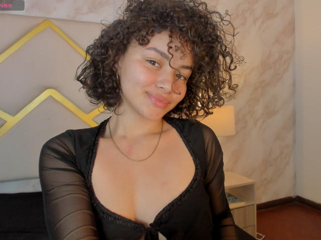 CurlyLorene, avec ses cheveux noirs bouclés, sa peau brun clair et son léger sourire, porte un haut noir transparent qui révèle ses petites seins et une chaîne en or. Décor : chambre à coucher..