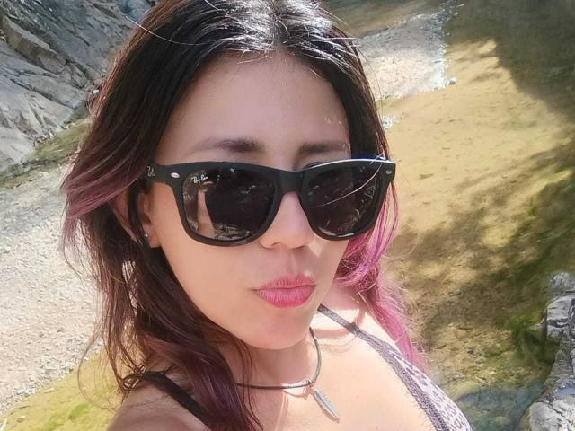 LunaTomson with pink-tipped brown hair, black cat-eye sunglasses, pink lipstick, and a black choker, showing off her tits in a sunlit outdoor setting.