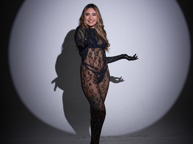 Ninilovee in piedi, con una body in pizzo nero che rivela il seno e i genitali, con lunghi capelli biondi, sorridente, indossa guanti neri, un'ombra su un muro bianco, corpo intero, sexy..