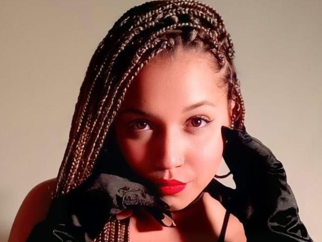 Athenea Clark, avec ses cheveux bruns tressés, son rouge à lèvres rouge, ses gants noirs, tenant son visage, regardant la caméra, lumière douce, expression sérieuse..