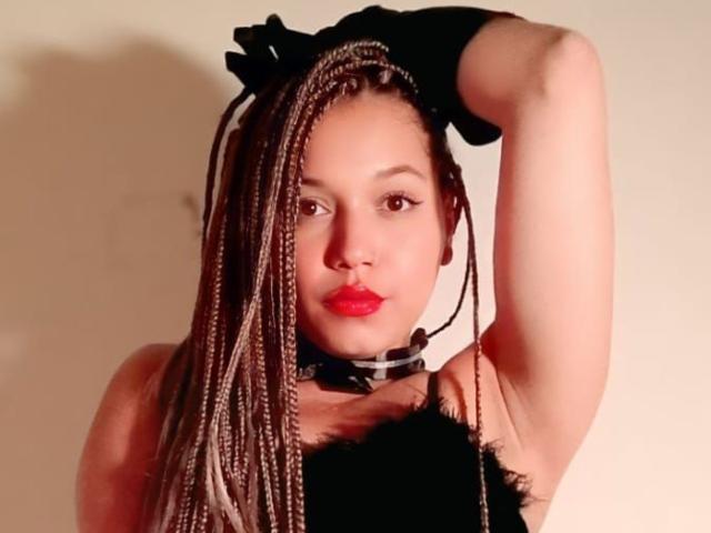 AtheneaClark with long braided hair, red lips, black gloves, and a black choker, raising her right arm, showing her armpit and black fur-covered chest.