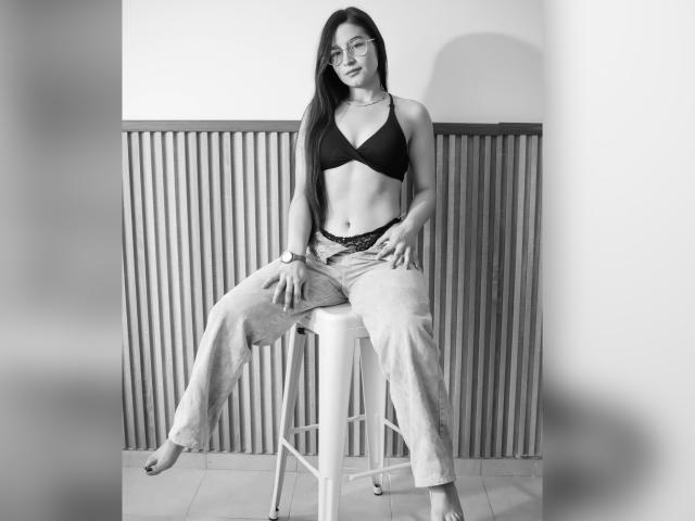 JadeHarpper sitting on a stool, wearing a black bra that shows her tits, lace panties, and loose jeans, with long dark hair, glasses, and a watch.