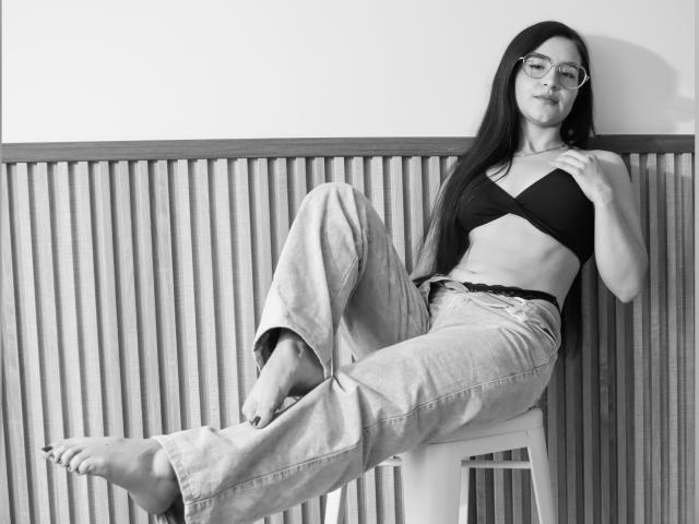 JadeHarpper with long dark hair, glasses, black bra, and loose jeans, legs up, leaning on chair, bare feet, looking at camera, casual, confident, monochrome.