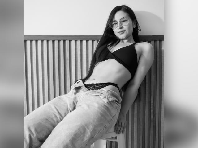 JadeHarpper with glasses, black bra, lace panties, and loose jeans, leaning against a wooden slat wall, smiling, showing a slim, toned body and a small, flat ass.