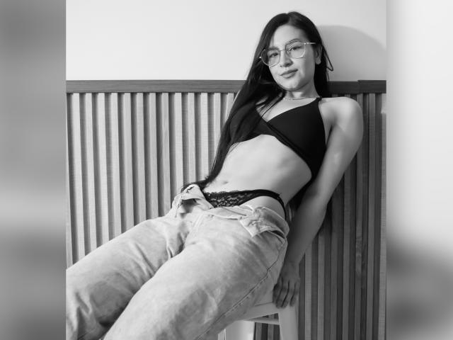 JadeHarpper leans back against a wooden railing, wearing glasses, a black bralette, and loose jeans with black lace panties showing. She has long dark hair and a slim build.