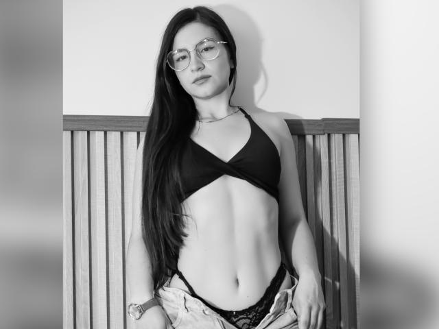 Black-and-white photo of JadeHarpper with long dark hair, glasses, black bra, lace panties, and unbuttoned jeans, showing a slim, flat-stomach, and small, perky tits.