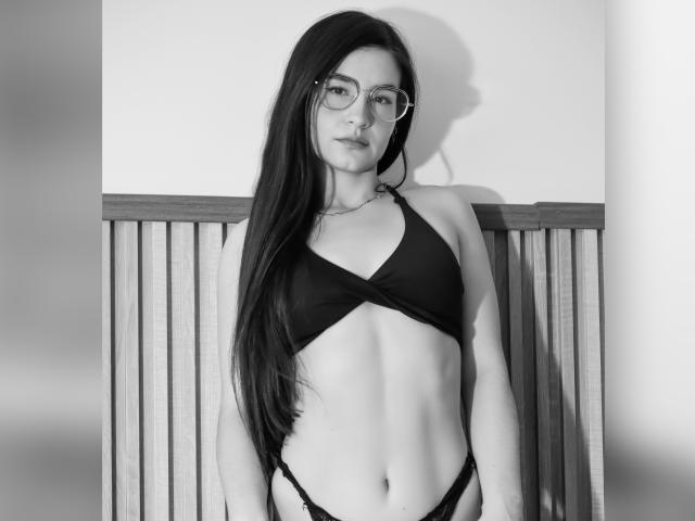 JadeHarpper with long black hair, glasses, black bikini top showing small tits, and black panties, standing against a wooden wall, black and white photo.