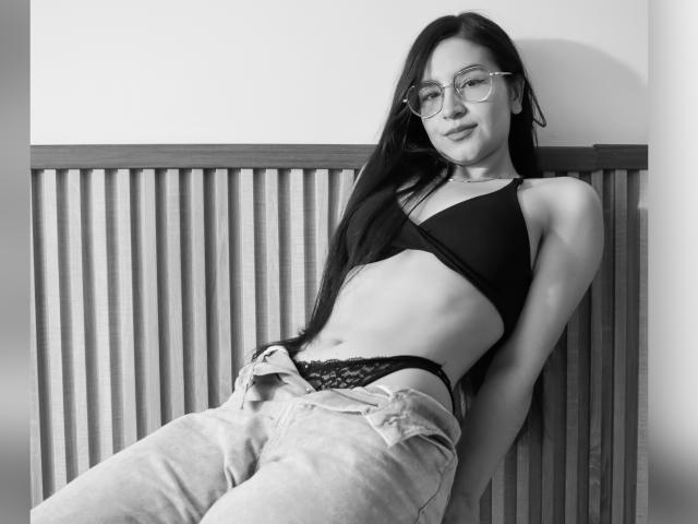 JadeHarpper with long black hair, glasses, black sports bra, showing a flat stomach and black lace panties, leaning against a wooden slat wall, wearing loose jeans.