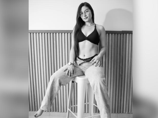 JadeHarpper with long dark hair, glasses, black bra, showing tits, and lace panties, sitting on stool, wearing loose jeans, hand on knee, black and white photo.