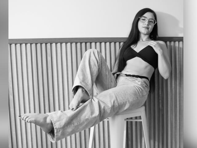 JadeHarpper with long dark hair, glasses, black bra, and loose jeans, sitting on a stool, legs up, bare feet, hand on chest, looking at camera, black-and-white.
