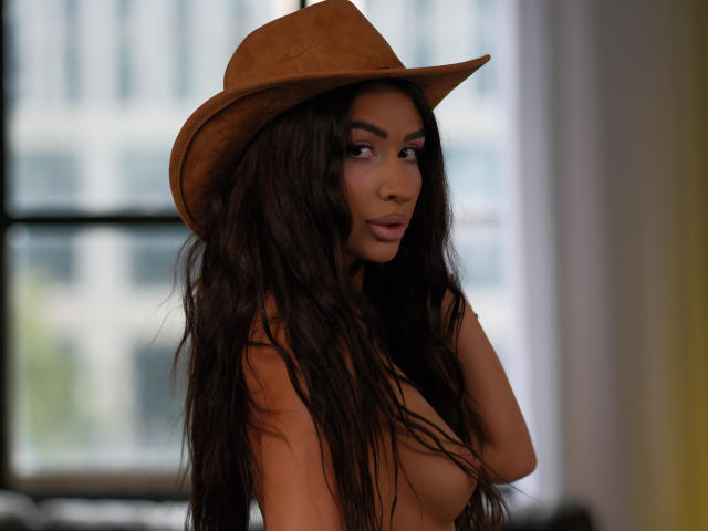 Emma Noor mit langem, gewelltem, dunklem Haar, die einen braunen Cowboyhut trägt, oben ohne, wobei kleine, pralle Brüste zu sehen sind, braune Augen und ein dezentes, selbstbewusstes Lächeln, in weichem, indirektem Licht..