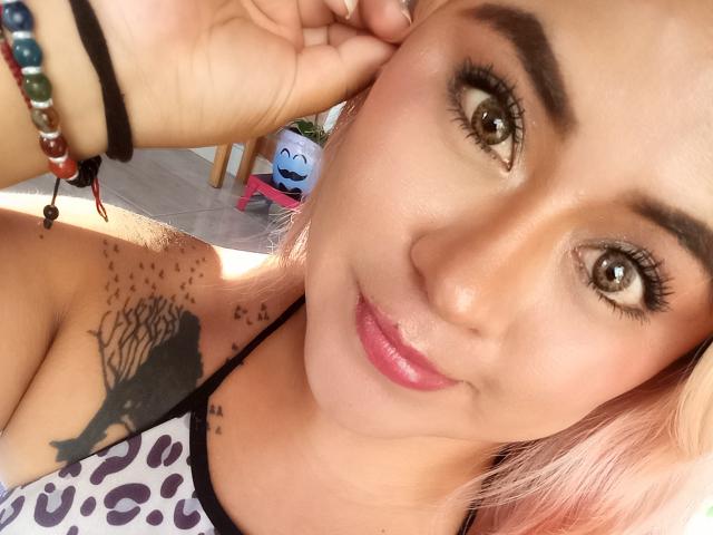 Estefaniaa69 with pink hair, brown eyes, and pink lipstick, showing a black tree tattoo on her shoulder, wearing a leopard-print top, and a beaded bracelet.