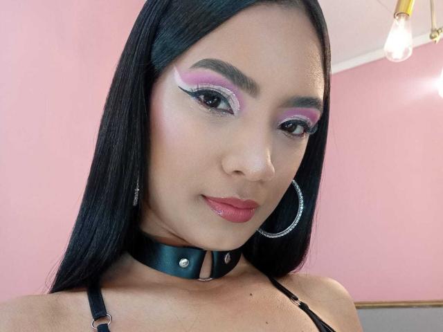 NatLewis with dark straight hair, pink and purple eyeshadow, red lipstick, black choker, hoop earrings, and black strappy top, showing a cute face with a slight smile.