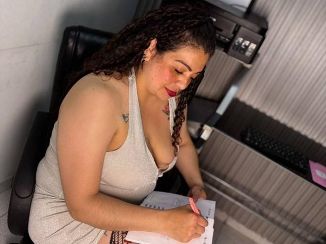 AlissaaMilf with curly brown hair, red lipstick, and a grey tank top, writing with a pink pen, showing her big tits and a small tattoo on her right shoulder.