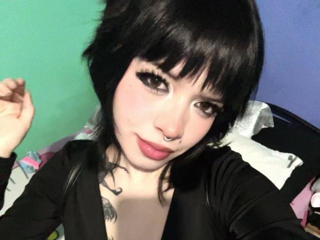 BabeSix with black hair, green eyes, and pink lips, wearing a black top, nose ring, and tattoo on her chest, smiling at the camera. Her tits are partially visible.
