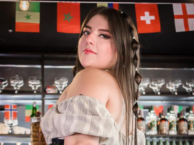 EmiilyStone, com longas tranças castanhas, blusa listrada com um ombro descoberto, olhando por cima do ombro, batom rosa, seios pequenos, pele clara, copos e garrafas de bebidas alcoólicas ao fundo..