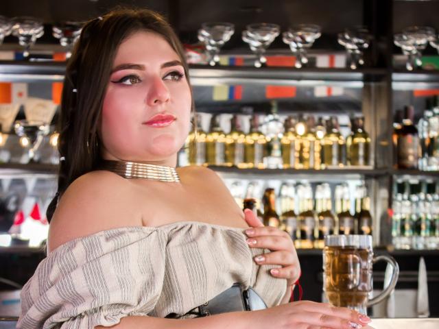 EmiilyStone, con il seno prosperoso, indossa una maglietta a righe con una spalla scoperta e una collana dorata, tiene in mano una boccale di birra, ha i capelli castano scuro, rossetto rosa e un'espressione leggermente compiaciuta..