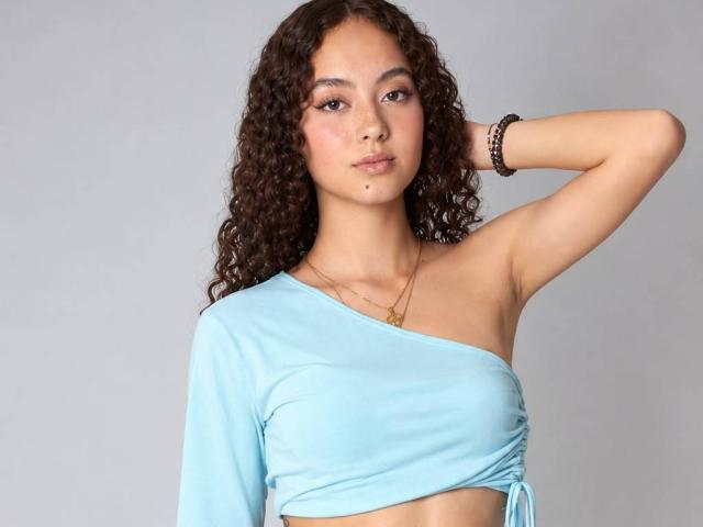 LeslyHoston with curly brown hair, wearing a light blue one-shoulder top, her small tits visible, arm raised, black bracelets, and a gold necklace, against a plain gray background.