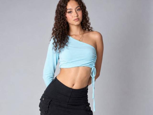 LeslyHoston has curly brown hair, wearing a light blue, one-shoulder crop top, exposing her small tits and black, high-waisted pants, standing with hands behind her back.