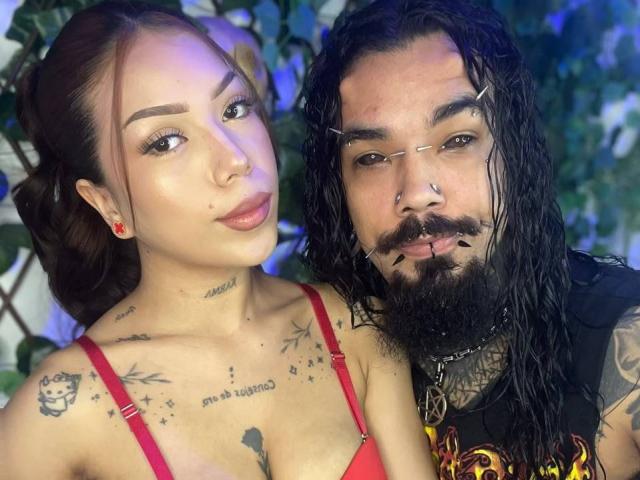 DiogenesYMaria with tats, red bra, and small tits, next to a bearded dude with wet hair, nose ring, and mustache, wearing a black shirt with fire designs.