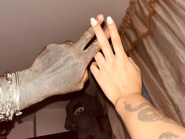 DiogenesYMaria's dark, wrinkled hand with silver bracelets holds DiogenesYMaria's lighter, tattooed hand with white nails. DiogenesYMaria's hand is on top, contrasting skin tones.