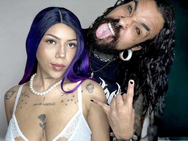 DiogenesYMaria with purple hair and white lace bra, showing tits, tattoos, and pearl necklace, standing next to a bearded man sticking out his tongue, with black hair and earrings.
