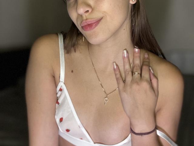 TrissyTerry, met een lichte huid, bruin haar en roze lippen, draagt een witte beha met rode hartjes, met haar hand op haar borst, een gouden ketting en ring, en glimlacht lichtjes. Borsten zichtbaar..