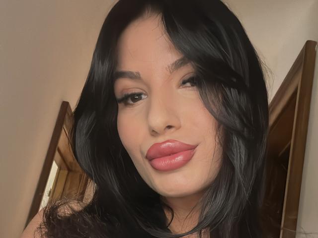 AmmyFrancaise with shiny black hair, winking, and pink glossy lips, wearing dark makeup, smiling subtly, showing off her smooth, fair-skinned face, and a hint of her upper chest.
