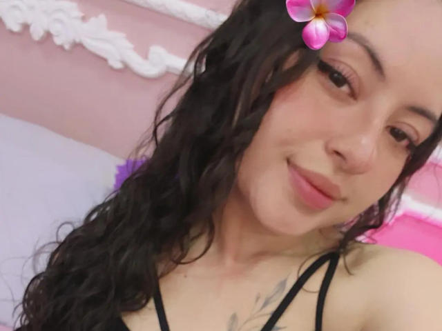 MiiaBennet hat welliges, schwarzes Haar, einen rosa Blumenfilter im Gesicht und ein leichtes Lächeln. Sie trägt ein schwarzes Oberteil, wobei ein Tattoo auf ihrer Brust sichtbar ist..