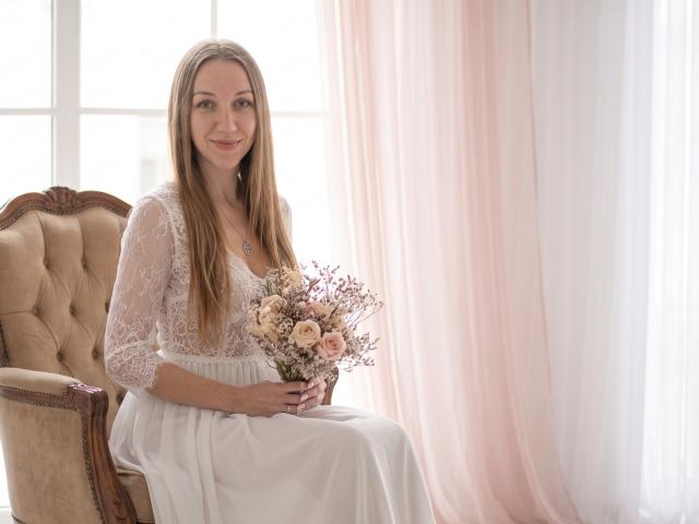 EllaMint, with long brown hair, lace top, and white dress, sits on a beige chair holding a bouquet of pink and white roses, smiling softly.