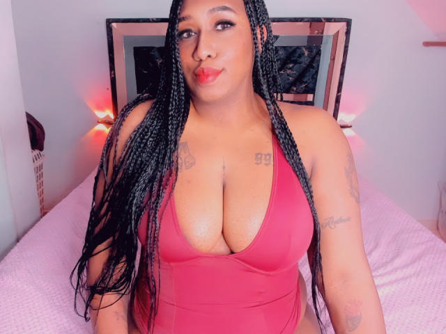Luxnyx with big tits, wearing a red one-piece, long braided hair, tattoos on chest and arm, pouty red lips, sitting on pink bed, pink lights in background.