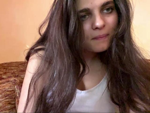 Jinny Kretschman, con lunghi capelli castano scuro, indossa una canotta bianca, mostra un sorriso appena accennato, ha la pelle chiara e gli occhi verdi. Il seno è leggermente visibile sotto la canotta..