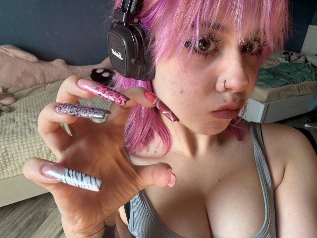 KrystalBerkovitch with pink hair, wearing headphones, grey tank top, showing off glittery pink and silver nails, holding out hand, nose ring, bedroom, light skin, large boobs.