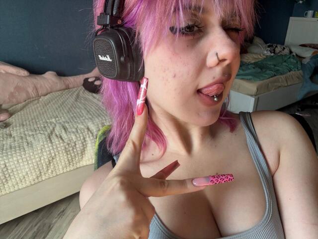 KrystalBerkovitch with pink hair, nose ring, and tongue piercing, wearing a headset, gray tank top, and pink nails, pointing at her cleavage, showing her tits.