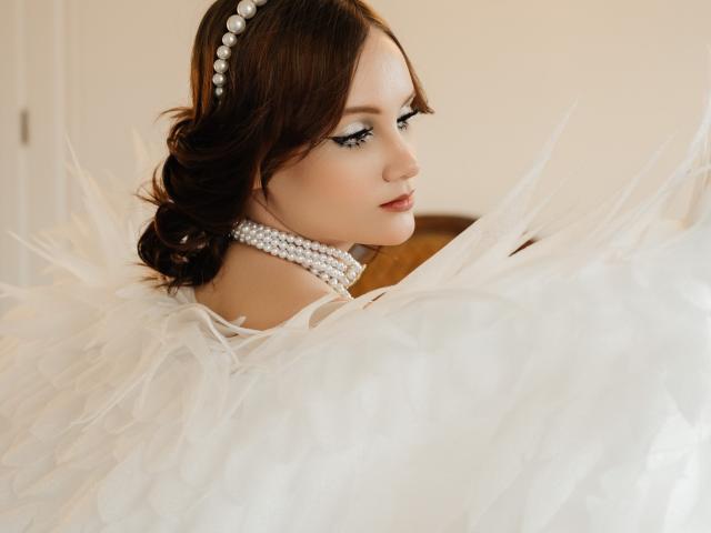 AngieBellas, con cabello castaño oscuro, vestida con un vestido blanco adornado con plumas, un collar de perlas y una diadema de perlas, mirando hacia un lado, con un maquillaje sutil, piel suave y una expresión elegante..