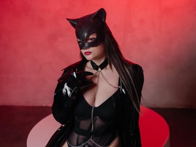 AngieBellas con una máscara de gato negro, cabello negro largo, lápiz labial rojo, un conjunto de látex negro, un collar de cadena y guantes, mostrando sus senos y sosteniendo una cadena contra su pecho..
