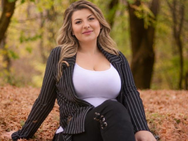 DalidaThierry with big boobs, wearing a pinstriped jacket over a white top and black pants, sitting on a leaf-covered ground, smiling, blonde wavy hair, forest background.