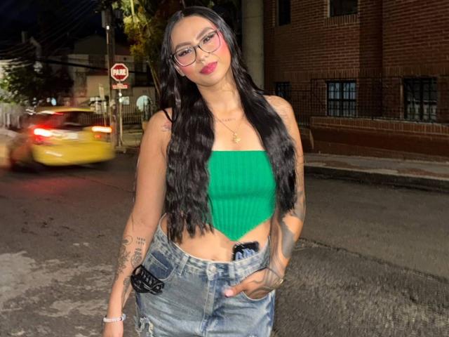 ChloeMoretti with long black hair, glasses, green tube top, and blue jeans, standing on a nighttime street, showing tattoos and a confident pose with a hand in her jeans.