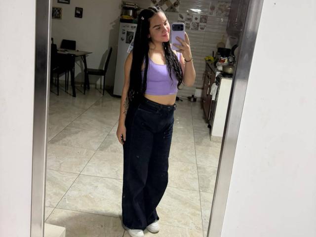 FenixAlthea with long black dreads, purple sports bra, showing small tits, and dark high-waisted jeans, taking a mirror selfie in a kitchen, wearing white sneakers.