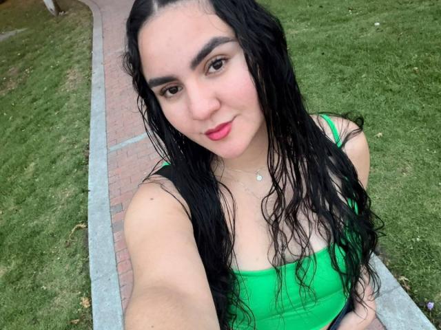 FenixAlthea has wet black hair, green tank top, and pink lipstick, showing off her small tits in a grassy area with a brick path. She's smiling at the camera.
