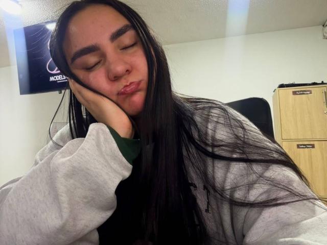 FenixAlthea with long black hair, eyes closed, pouting lips, hand on cheek, wearing a gray hoodie, sitting in a chair, looking relaxed and cute.