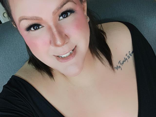GiiannaMorrys has a chubby face, black hair, and a tattoo on her shoulder that reads "My Future's". She's smiling with pink cheeks, wearing a black top, and earrings.