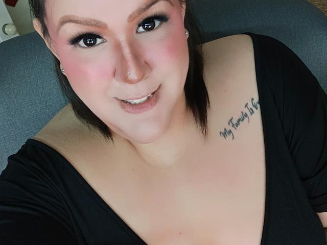 GiiannaMorrys has fair skin, black hair, and is smiling with pink blush on her cheeks, wearing a black top, showing a tattoo on her right shoulder that reads "My favorite thing."