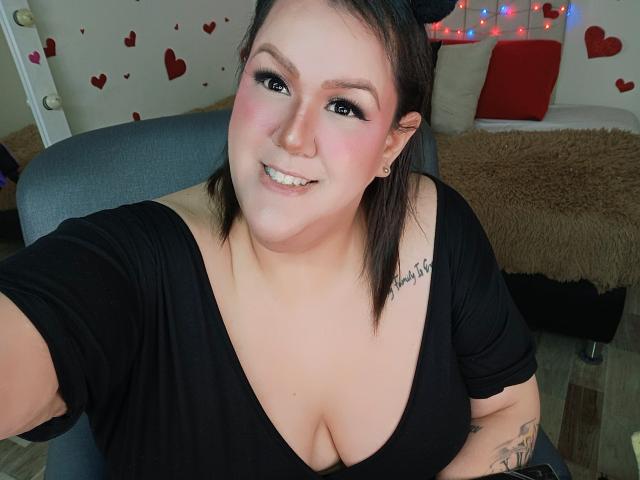 GiiannaMorrys, chubby with pale skin, black hair, wearing a black top, showing big tits, smiling, black eyeliner, tattoo on right shoulder, cat ears, wooden floor, chair.