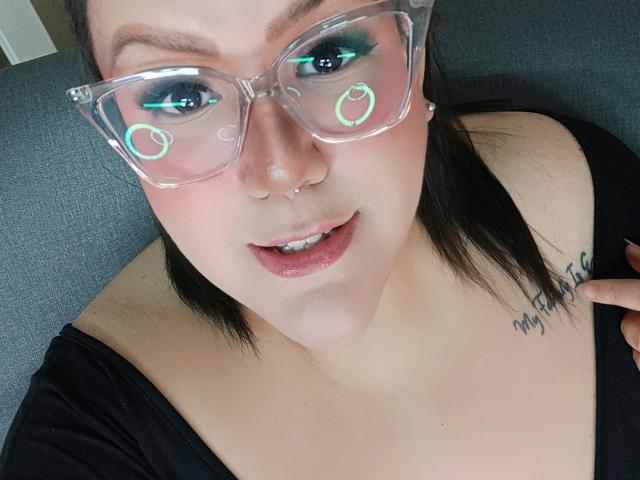 GiiannaMorrys with clear glasses, teal eyeliner, and a nose ring, showing her slightly open mouth and black tattoo on her right shoulder, wearing a black top.