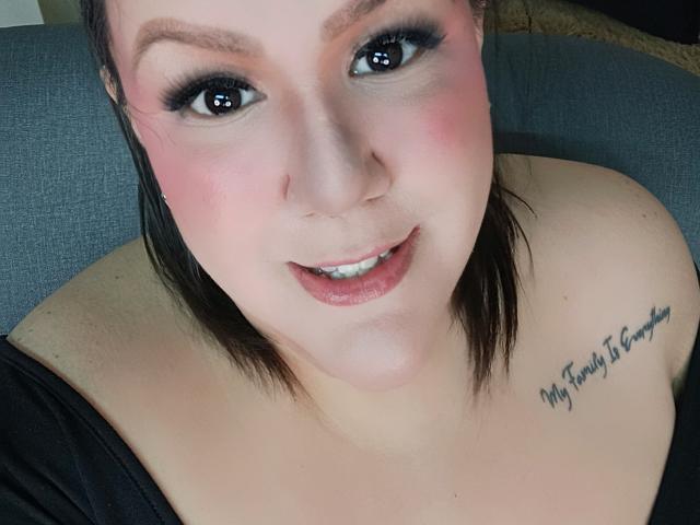 GiiannaMorrys has pink cheeks, black eyeliner, and a tattoo on her right shoulder that reads "My Family Is Everything". She's looking up with a smile, showing her tits.