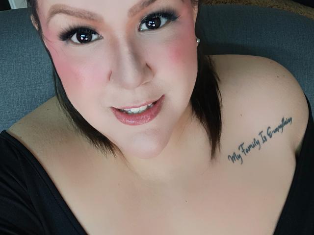 GiiannaMorrys with pink blush, black eyeliner, shiny pink lips, black top, visible tattoo on right shoulder saying "My family is everything." Her tits are partially visible.
