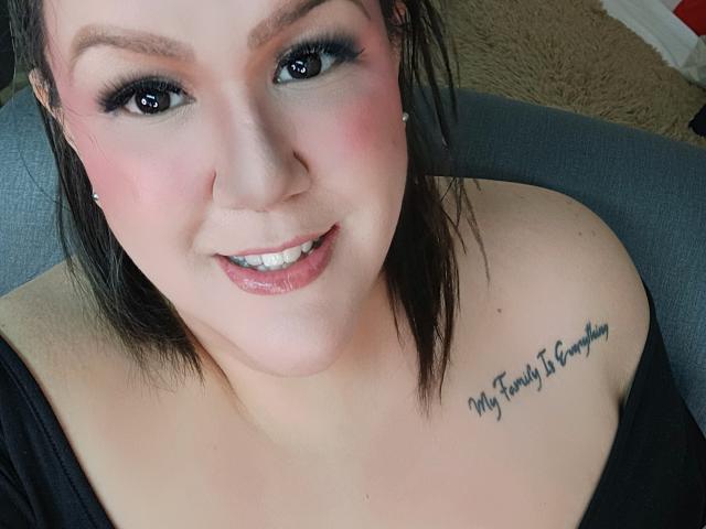 GiiannaMorrys has pink-tinted skin, black eyeliner, and a tattoo on her right shoulder that reads "My Family Is Everything." She's smiling with glossy lips and black top.