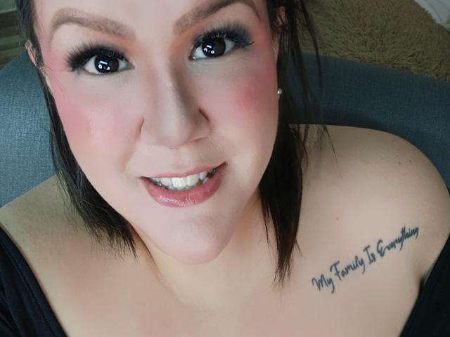 GiiannaMorrys with pink blush, black eyeliner, and glossy pink lips, showing her bare shoulders and tattoo on her right boob that reads "My Family Is Everything."