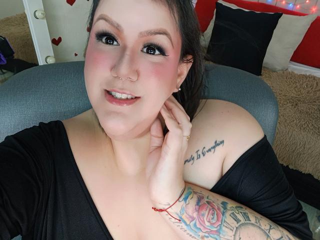 GiiannaMorrys with black eye makeup, pink blush, nose ring, smiling, black off-shoulder top, left hand touching neck, tattoo on right shoulder, colorful tattoo on right arm, chair.